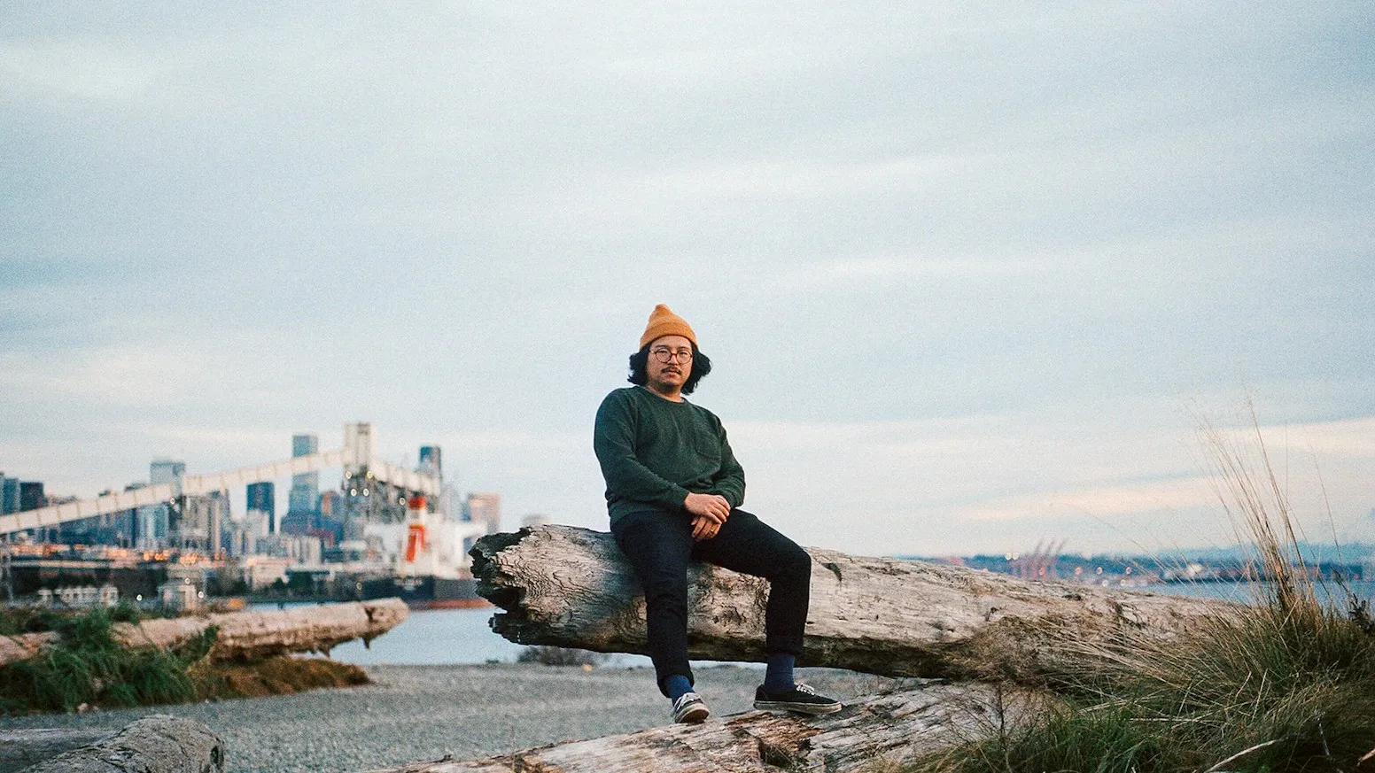 The musician Byung sitting on a driftwood log by the water, with a city skyline behind him.