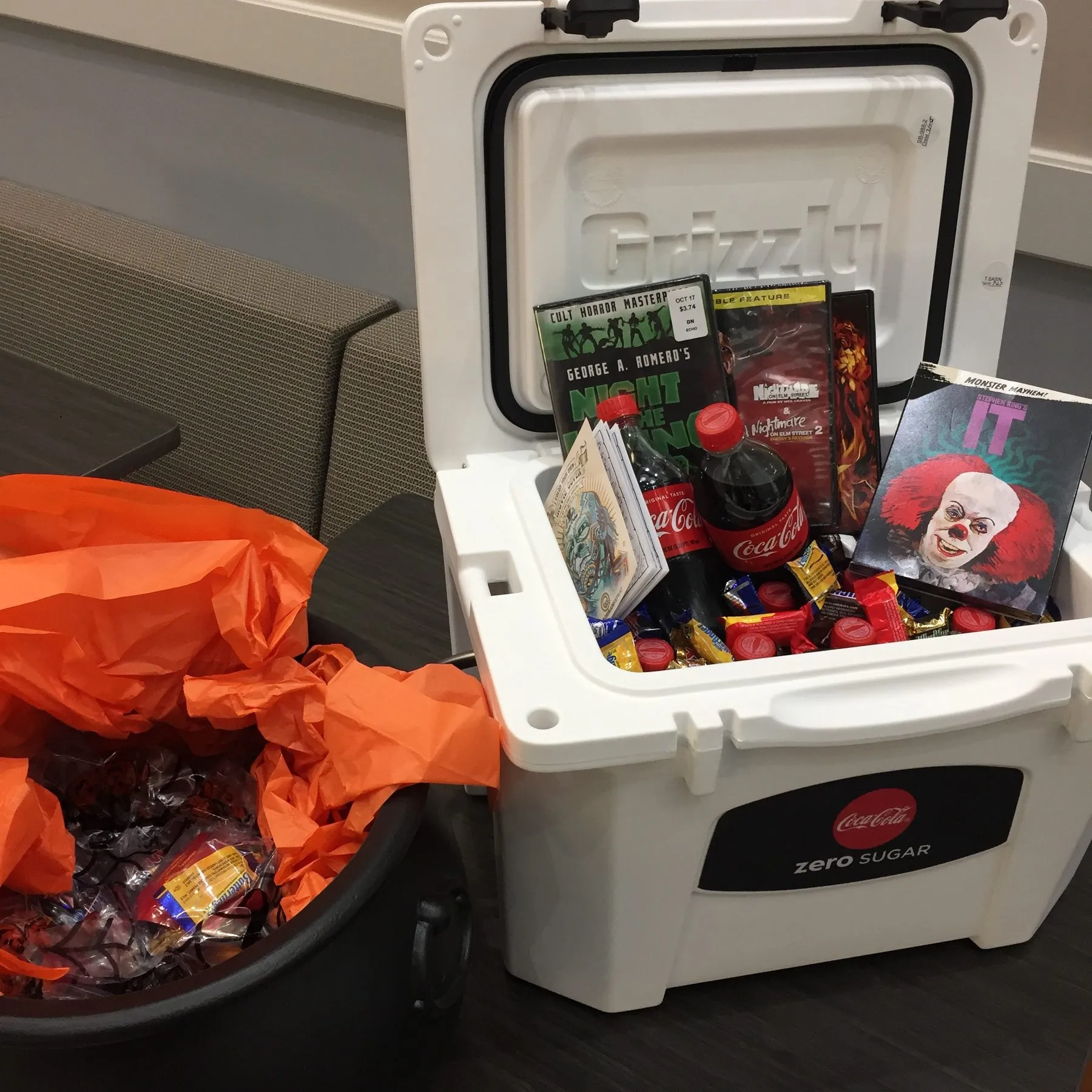 A cooler of Halloween prizes, including Coca-Cola, candy, and an IT movie cover, next to a bin of wrapped candy.