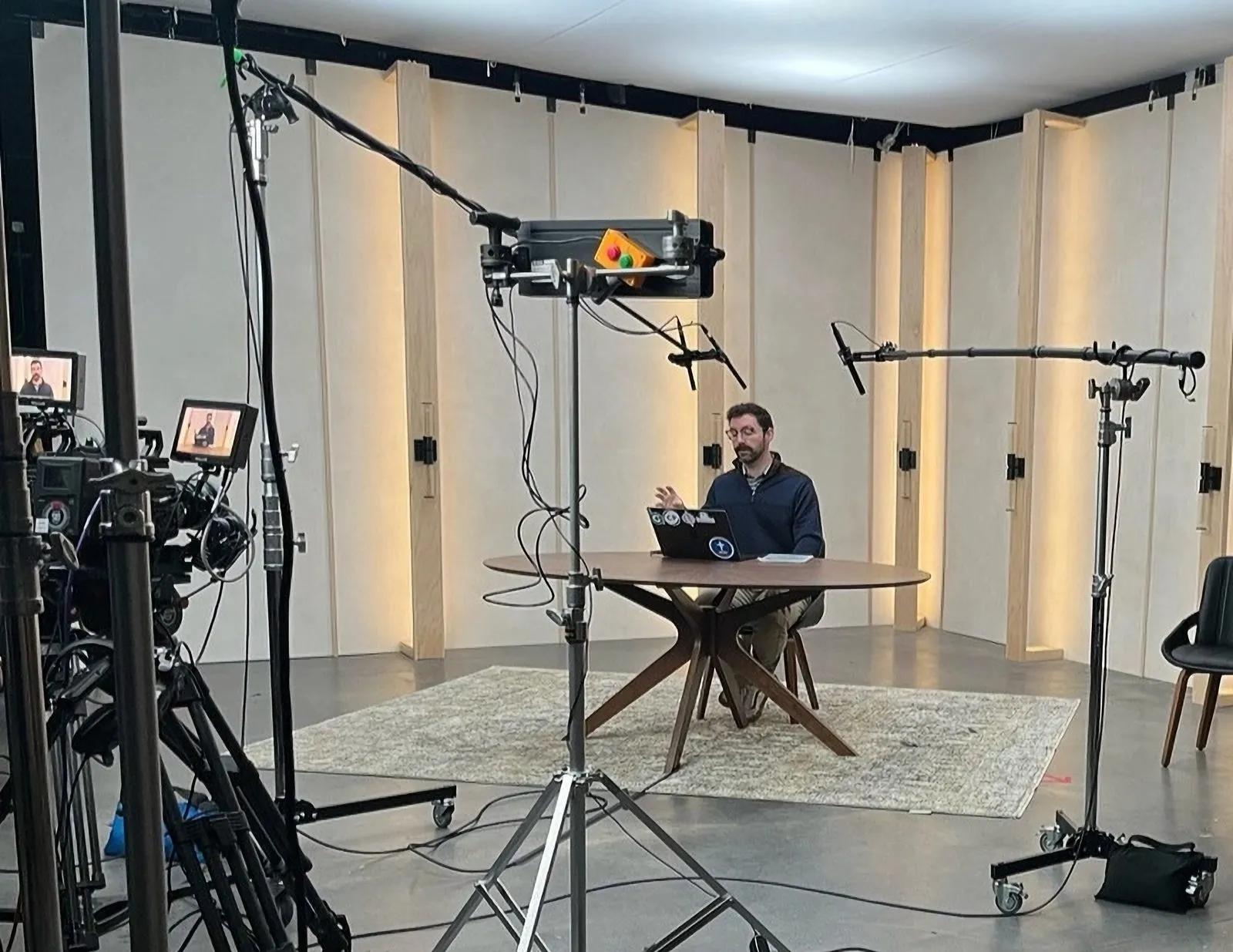 Mitchell at a table in a video studio, surrounded by cameras, lights, and microphones.