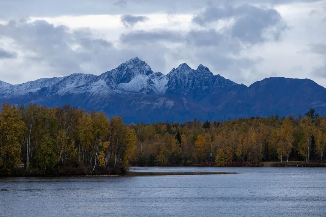 Snow-dusted mountains above a lake lined with autumn trees.