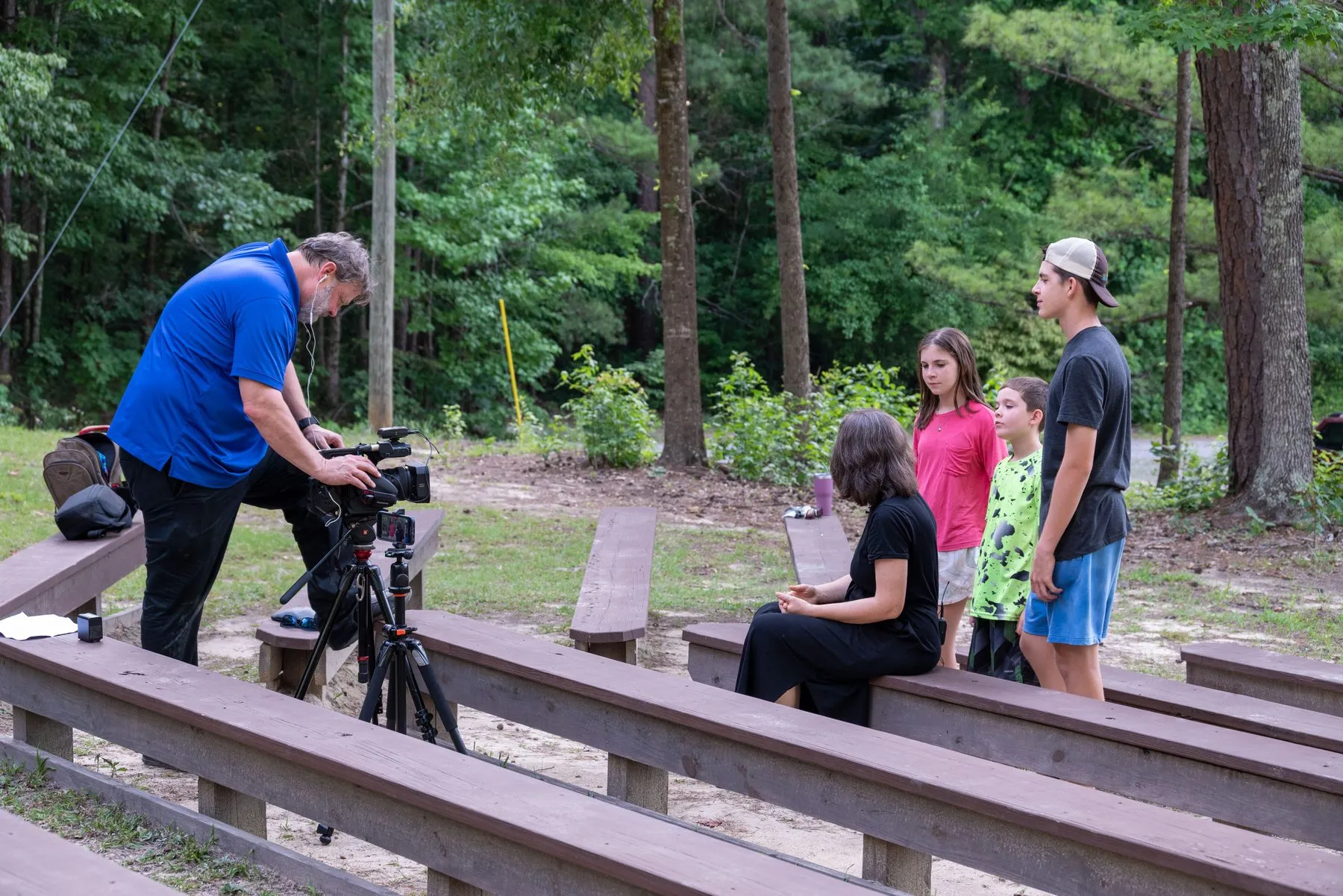 A camera operator filming people at an outdoor pavilion in the woods.