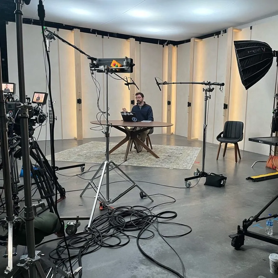 A video studio with lights and cameras set up around a man at a round table.