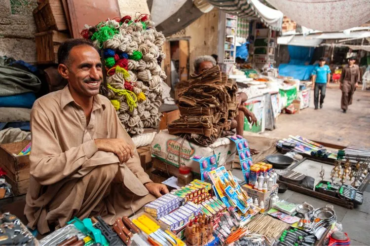 A smiling street vendor seated among colorful goods in a busy outdoor market.